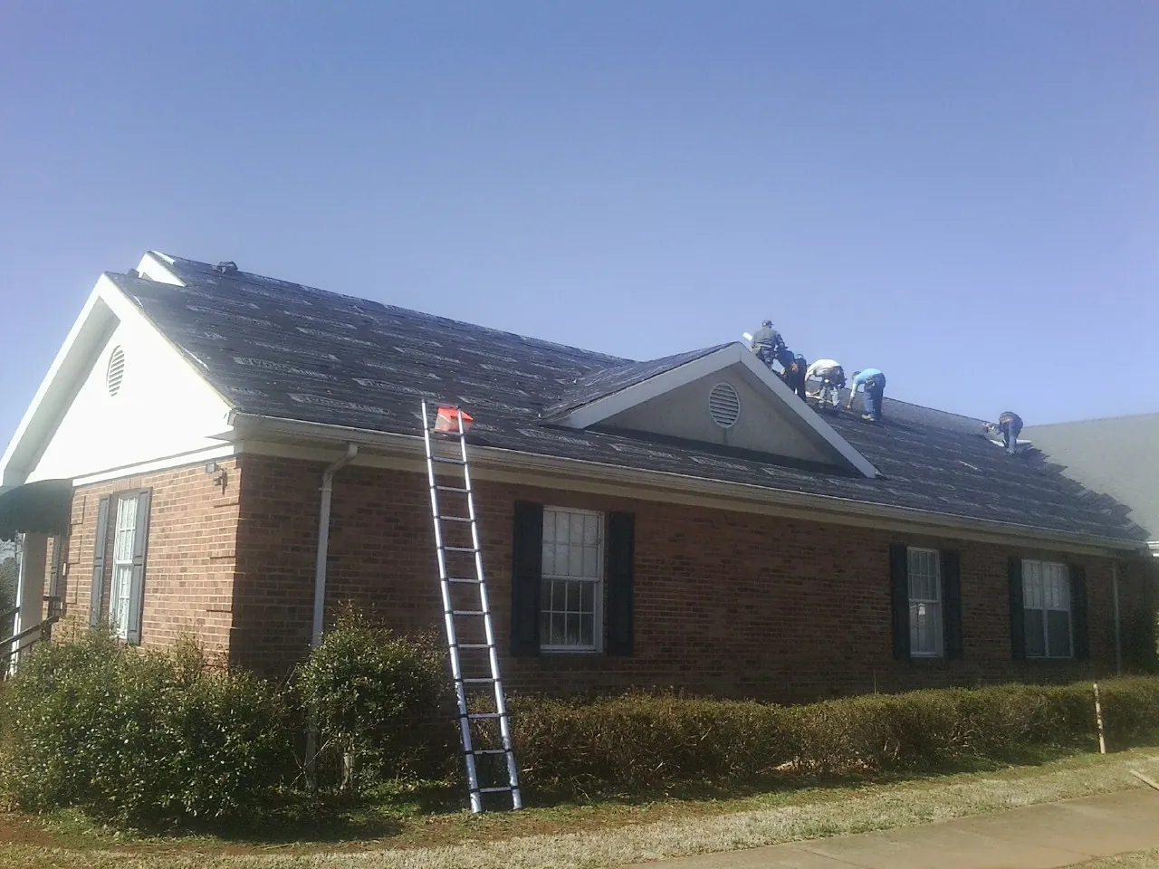 Crew tearing shingles off a brick ranch roof with a ladder set — Metal Roof Repair work by our crew in Pheba
