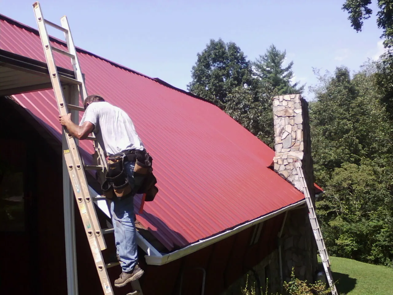 Roofer on a ladder beside a red metal roof and stone chimney — Soffit & Fascia Repair by Roofing Brothers in Pheba